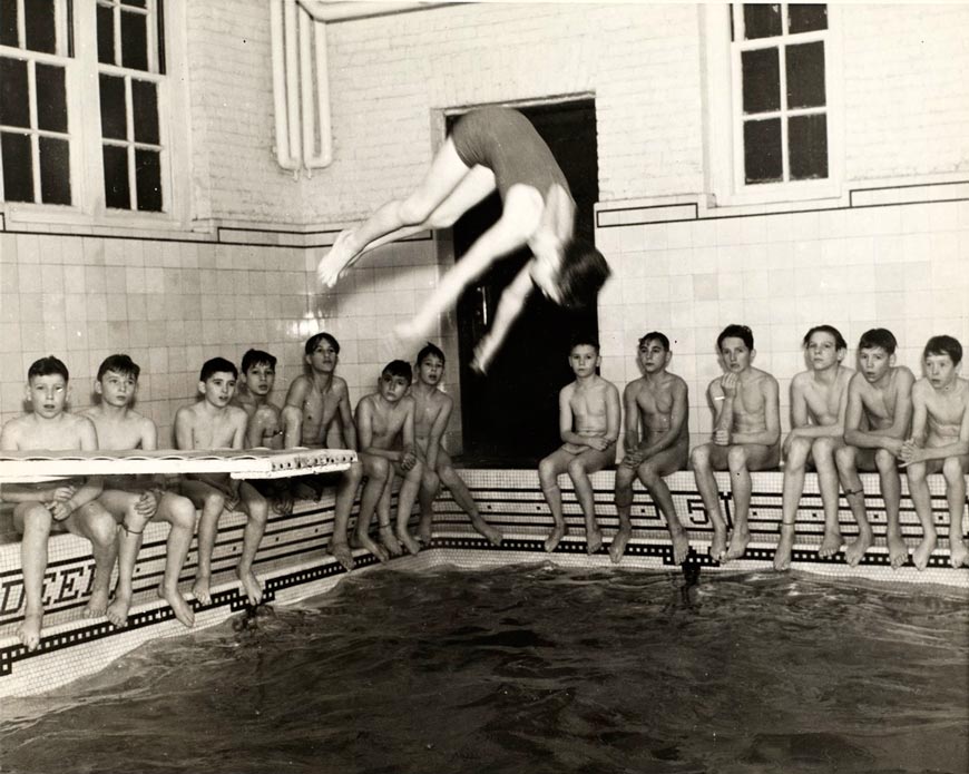 A boy performs a dive in a natatorium pool as a group of boys look on (Мальчик совершает прыжок в бассейн, а группа мальчиков наблюдает)