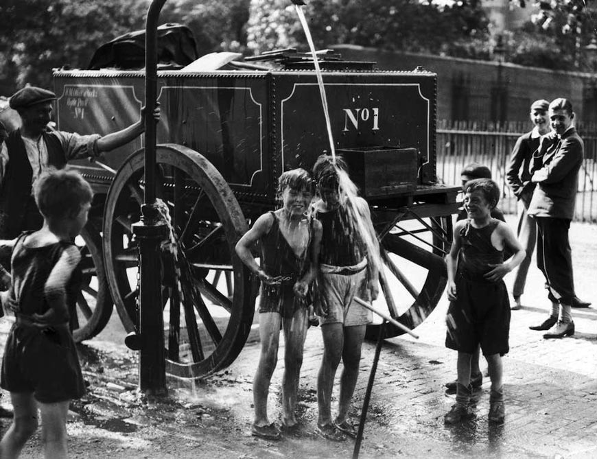 A water cart man turns the water main on a group of boys to help them cool off in a street (Мужчина водовоза направляет водовод на мальчишек, помогая им охладиться на улице), August 1930