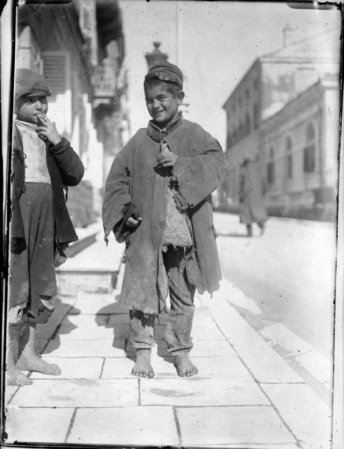 Boy smoking a cigarette next to a smiling refugee on the street, wearing a soldier's coat, a piece of a bedquilt and pajama trousers (Мальчик курит сигарету рядом с улыбающимся беженцем на улице, одетым в солдатскую куртку, кусок простыни и пижамные штаны), 1919