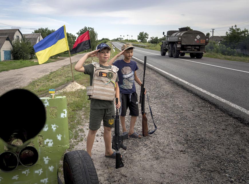 Максим и Андрей [11], отдают честь украинским солдатам, держа пластиковое оружие, играя на самодельном блокпосту на трассе (Maksym and Andrii [11], salute to Ukrainian soldiers holding plastic guns as they play at the self-made checkpoint on the highway), 2022