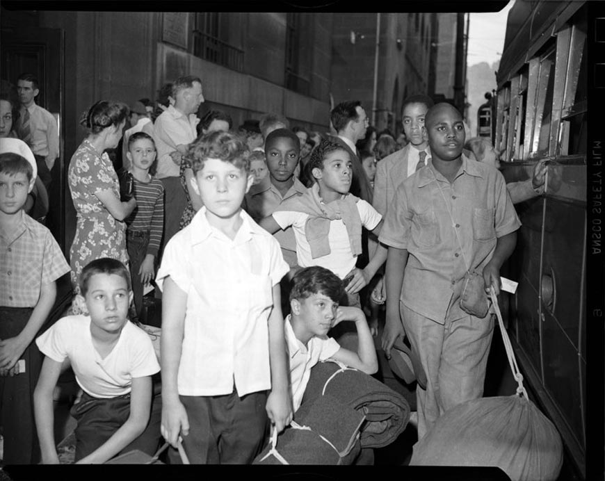 Boys holding blankets and sleeping bags, with men and women in background, on sidewalk with bus on right (Мальчики держат одеяла и спальные мешки, на заднем плане мужчины и женщины на тротуаре и автобус справа), 1940-1945