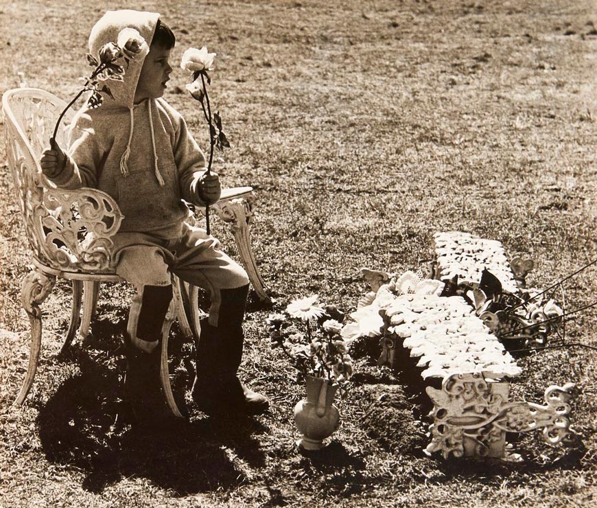 Untitled / A young boy in a hooded sweat suit and boots seated outside and holding flowers (Без названия / Мальчик в спортивном костюме с капюшоном и ботинках сидит на открытом воздухе и держит цветы)