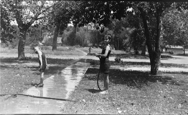 Boy spraying smaller boy with water from hose, surrounded by grass and trees (Мальчик обливает мальчика по-младше водой, на газоне под деревьями), c.1950s