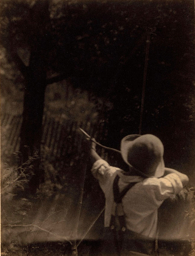 [Back view of boy in hat and overalls, aiming homemade bow and arrow, with tree in background (Вид сзади мальчика в шляпе и комбинезоне, целящегося из самодельного лука со стрелой, на фоне дерева)]