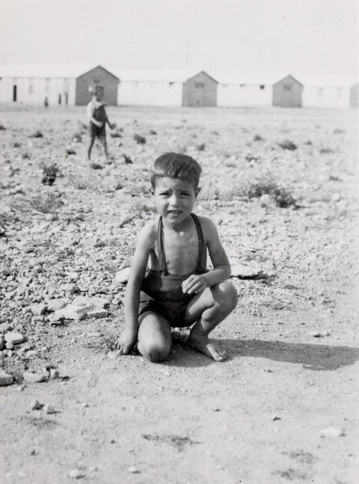 A small boy [probably Spanish] crouches down, as he plays in the dirt in front of the barracks of the Rivesaltes internment camp (Маленький мальчик [вероятно, испанец] присел на корточки, играя в грязи перед бараками лагеря для интернированных Ривесальтес), c.1942
