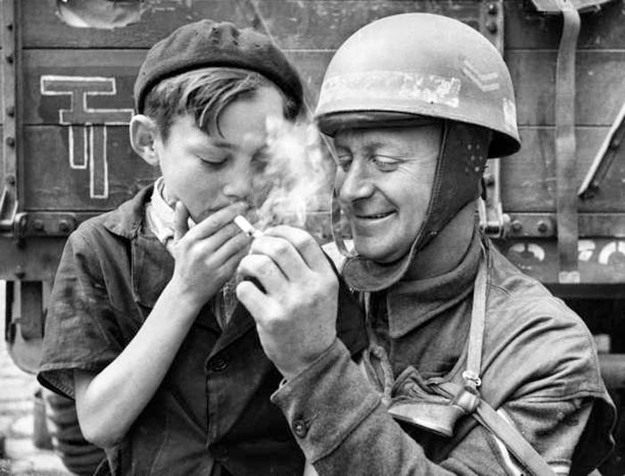 Corporal Fred Sicklong, a motorcycle despatch rider from XXX Corps, British 2nd Army lights a cigarette for a young French boy in the liberated town of Bayeux following Operation Overlord, the D-Day Allied invasion of German-occupied France (Капрал Фред Сиклонг, мотострелок 30-го корпуса 2-й британской армии, прикуривает сигарету французскому мальчику в освобожденном городе Байё после операции «Оверлорд» — вторжения союзников в оккупированную немцами Францию ​​в день «Д»), 1944