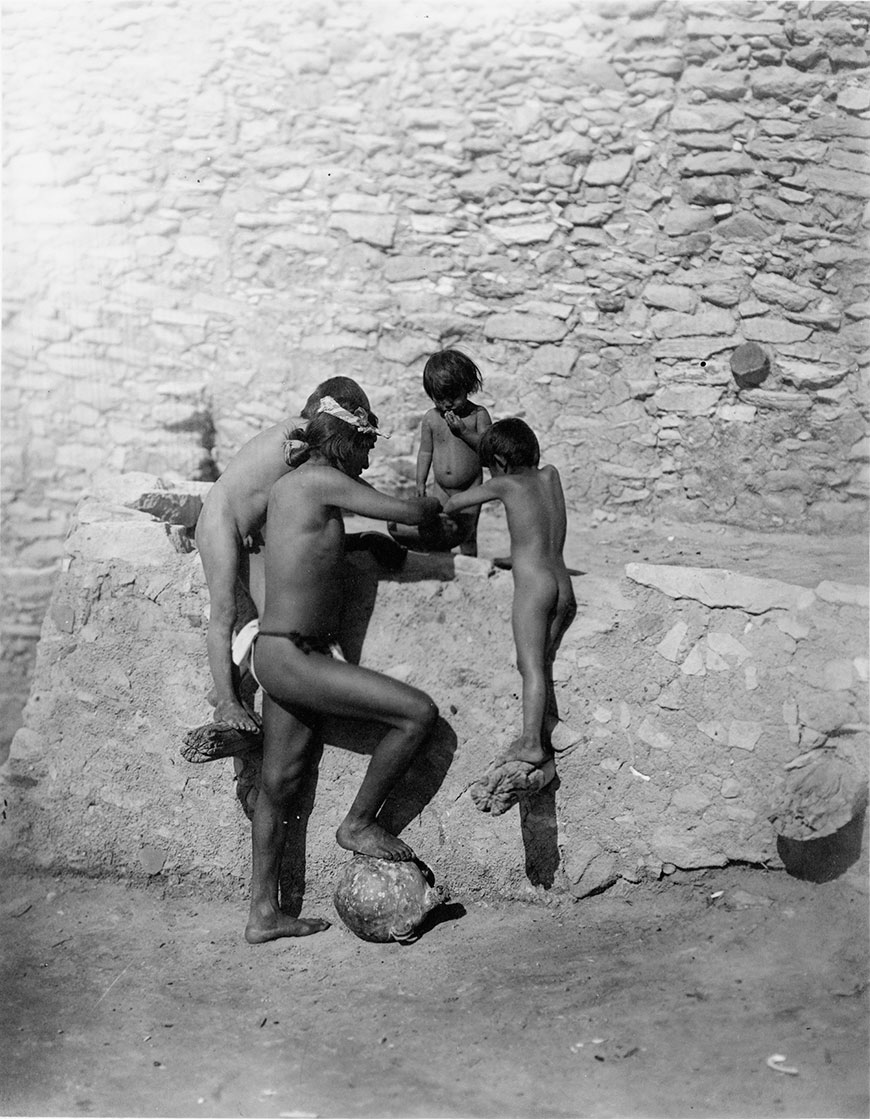 Four Hopi Indians (1 adult and 3 children), wearing little or no clothing eating out of bowl next to stone structure (Четверо индейцев хопи, взрослый и три ребёнка, практически без одежды, едят из чаши рядом с конструкцией из камня), c.1913